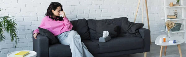 病気の若い女性がソファに座って生活室で紙ナプキンで鼻を吹いて バナー — ストック写真