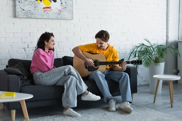 Sorrindo Jovem Tocando Guitarra Acústica Perto Namorada Sentada Sofá Casa — Fotografia de Stock