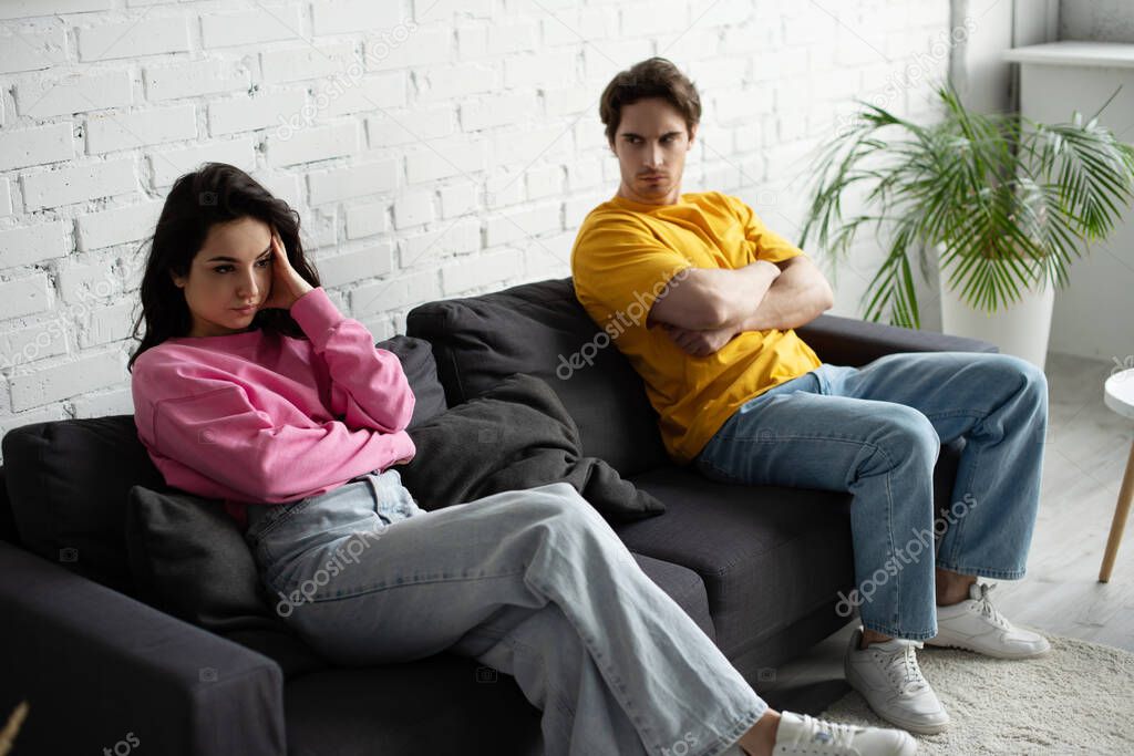 angry young man with crossed arms sitting on couch and looking at girlfriend with hand near head in living room