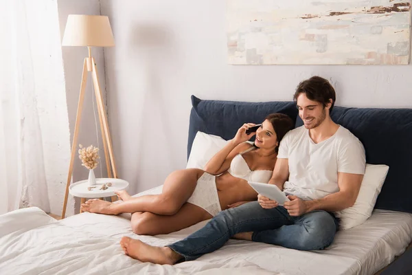 ベッドルームにデジタルタブレットを持つボーイフレンドの近くのスマートフォンで話す下着姿の笑顔の女性 — ストック写真