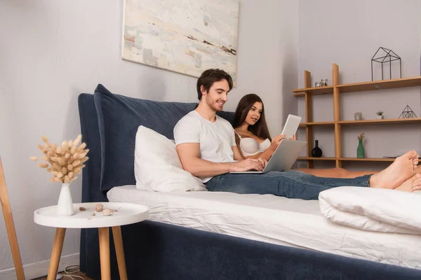 Sorrindo Homem Digitando Laptop Perto Mulher Lingerie Usando Smartphone Quarto — Fotografia de Stock