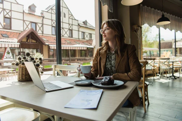 Young Freelancer Cup Looking Away Cake Laptop Menu Cafe — Stock Photo, Image