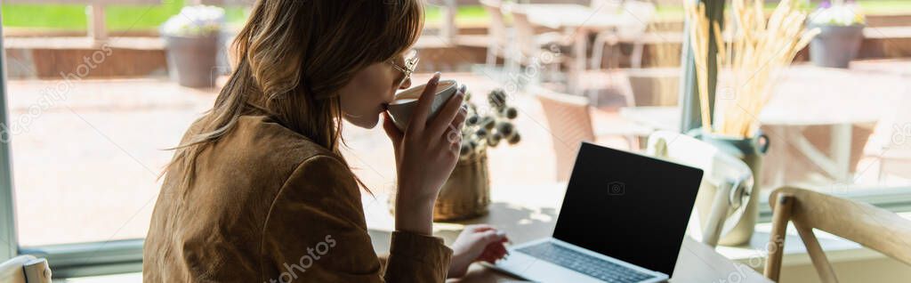 Side view of young woman dinking coffee near blurred laptop in cafe, banner 