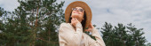 Tiefansicht einer fröhlichen jungen Frau mit Sonnenbrille und Strohhut in Waldnähe, Banner