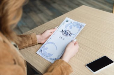 Kafedeki boş ekranlı cep telefonunun yanında menüyü tutan kadının kırpılmış görüntüsü 