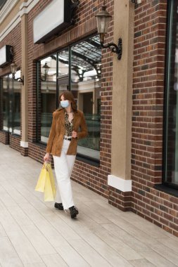 Medikal maskeli, alışveriş merkezinin yakınında alışveriş çantalarıyla yürüyen bir kadın.