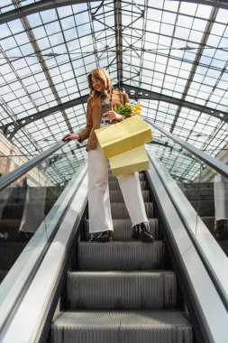 full length of happy woman in sunglasses holding yellow tulips and shopping bags while standing on escalator  clipart