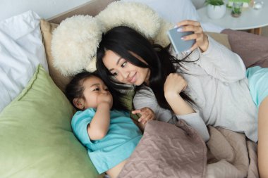 Akıllı telefonlu gülümseyen anne yatakta Asyalı çocuğa bakıyor. 