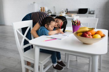 Asyalı ebeveyn kızını öpüyor ve evde defter çiziyor. 
