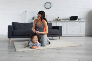 Spor kıyafetli Asyalı anne elinde tokayla gülümseyen çocuğun yanında. 