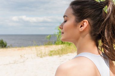 Genç ve esmer bir kadın, gözleri kapalı, okyanus kenarında meditasyon yapıyor.