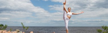 Spor kıyafetleri içinde neşeli bir kadın. Baş parmak, deniz kenarında poz veriyor.