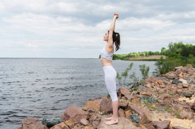Spor kıyafetli genç bir kadın deniz kenarında yoga yapıyor.