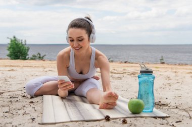 Kulaklıklı gülümseyen kadın yoga minderine uzanırken müzik dinliyor ve akıllı telefon kullanıyor. 
