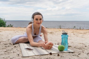 Kulaklıklı neşeli kadın yoga minderine uzanırken müzik dinliyor ve akıllı telefon kullanıyor. 