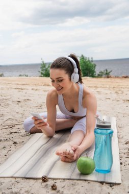 Kablosuz kulaklıklı gülümseyen kadın yoga minderine uzanırken müzik dinliyor ve akıllı telefon kullanıyor. 