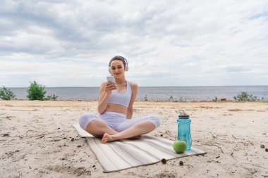 Kablosuz kulaklıklı gülümseyen kadın akıllı telefon kullanırken müzik dinliyor ve yoga minderinde bacak bacak bacak üstüne atmış bir şekilde oturuyor. 