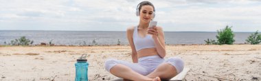 Kulaklıklı gülümseyen kadın akıllı telefon kullanırken müzik dinliyor ve yoga minderinde bacak bacak üstüne atmış bir şekilde oturuyor.