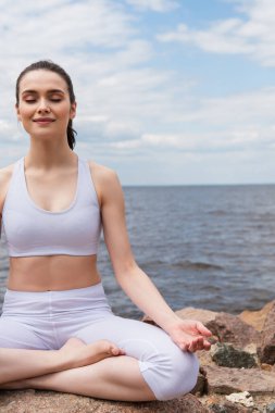 Spor kıyafetli neşeli kadın deniz kenarında meditasyon yaparken lotus pozu veriyor. 