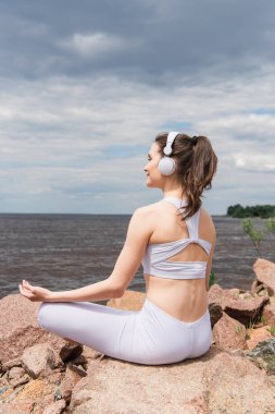 Mutlu kadın kulaklıklar ve spor kıyafetleriyle deniz kenarında meditasyon yaparken lotus pozu veriyor. 
