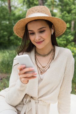 Hasır şapkalı ve kablosuz kulaklıklı mutlu genç kadın ormanda bulanık akıllı telefon kullanıyor. 