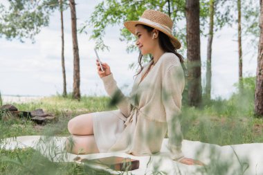 Piknik battaniyesinde otururken akıllı telefon kullanan hasır şapkalı ve kablosuz kulaklıklı bir kadın. 