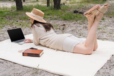 Hasır şapkalı ve kulaklıklı kadın akıllı telefon, güneş gözlüğü ve kitabın yanındaki piknik battaniyesinde yatarken boş ekranlı laptop kullanıyor. 