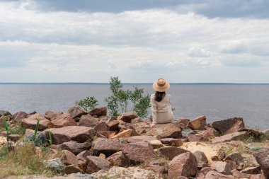 Hasır şapkalı, denizin kenarında oturan genç bir kadının arka görüntüsü.