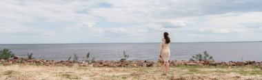 Uzun boylu, esmer, genç bir kadın elinde güneş şapkası ve denizin yanında duruyor.