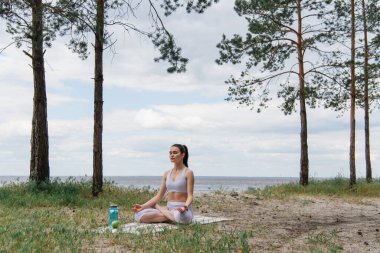 Spor kıyafetli esmer bir kadın lotus poz verip yoga minderinde meditasyon yapıyor. 