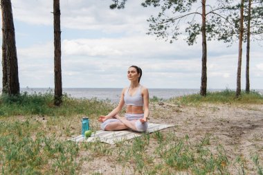 Spor kıyafetli esmer kadın lotus poz verip yoga minderinde meditasyon yapıyor. 