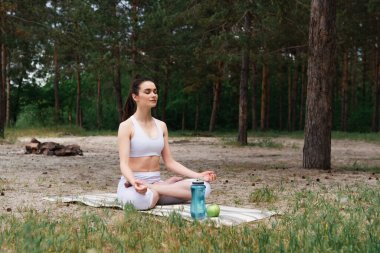 Spor kıyafetli barışçıl genç bir kadın lotus poz verip yoga minderinde meditasyon yapıyor. 