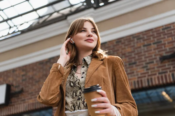 low angle view of pleased woman in wireless earphones holding paper cup and adjusting hair outside