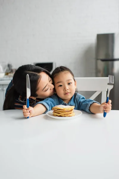 Asiática Madre Besos Niño Con Cubiertos Cerca Panqueques Mesa — Foto de Stock