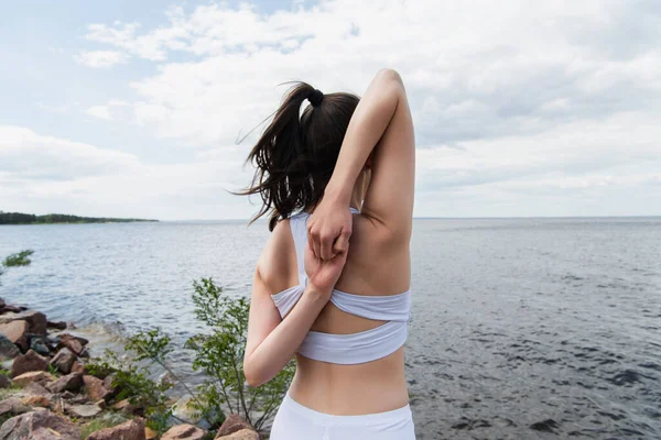 back view of young woman doing cow face pose with hands behind back near sea