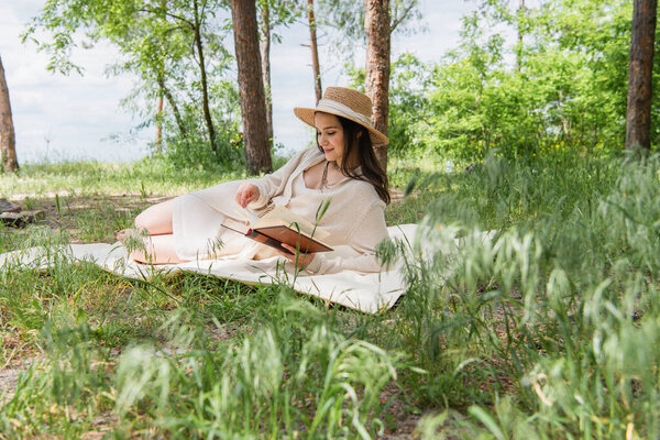 joyful young woman in straw hat reading book in forest 