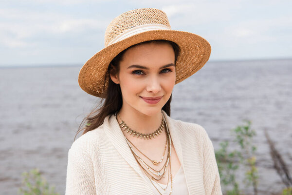 pleased young woman in sun hat looking at camera near sea