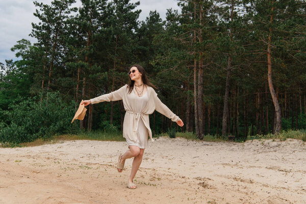 full length of positive young woman in sunglasses holding straw hat and walking near forest 