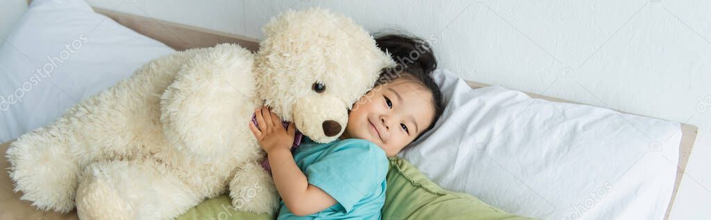 Asian child lying with teddy bear on bed, banner 