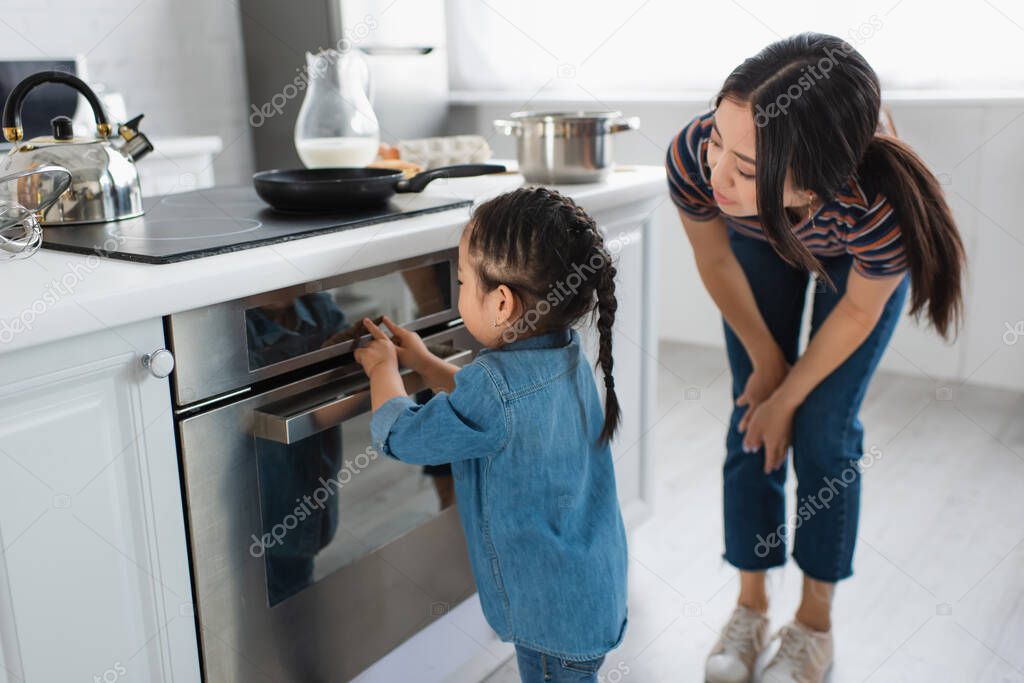 Asian kid touching display of oven near mother in kitchen 