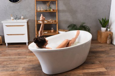 Çağdaş küvette elini kaldırmış neşeli genç bir kadın banyo yapıyor. 
