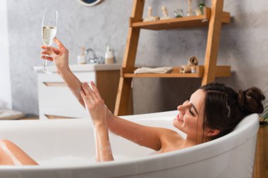 Küvette banyo yaparken elinde şampanya bardağıyla mutlu bir kadın.