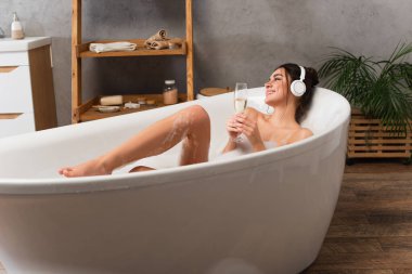 pleased woman in headphones listening music and holding glass of champagne while taking bath  clipart