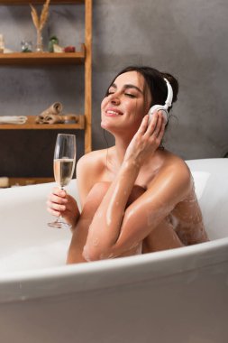 Kulaklıklı neşeli bir kadın müzik dinliyor ve banyo yaparken elinde bir bardak şampanya tutuyor. 
