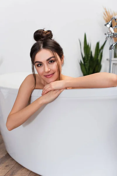 happy young woman with hair bun looking at camera in bathtub