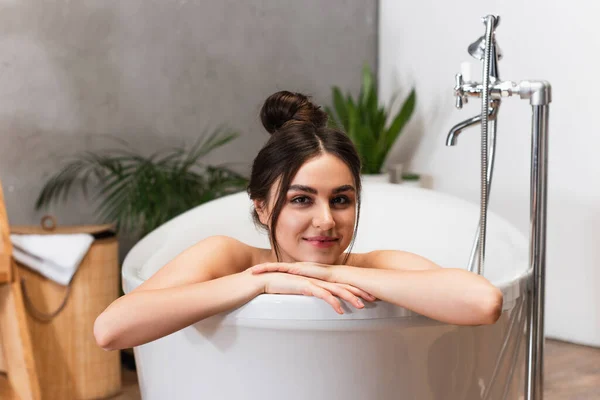 joyful young woman with hair bun looking at camera in bathtub