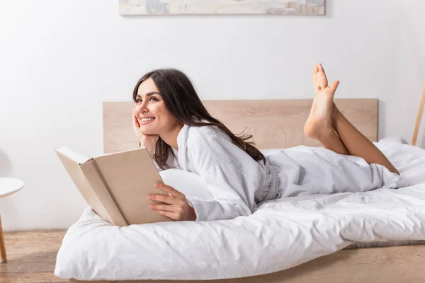 穿着浴衣的快乐女人躺在床上看书 — 图库照片