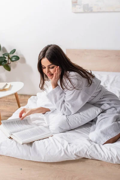 Mulher Morena Roupão Sentado Cama Sorrindo Enquanto Livro — Fotografia de Stock