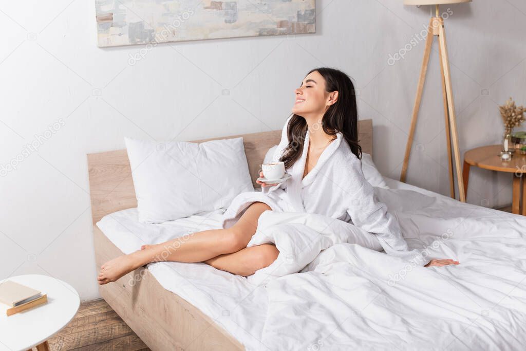 happy young woman holding cup of coffee and saucer in bedroom 