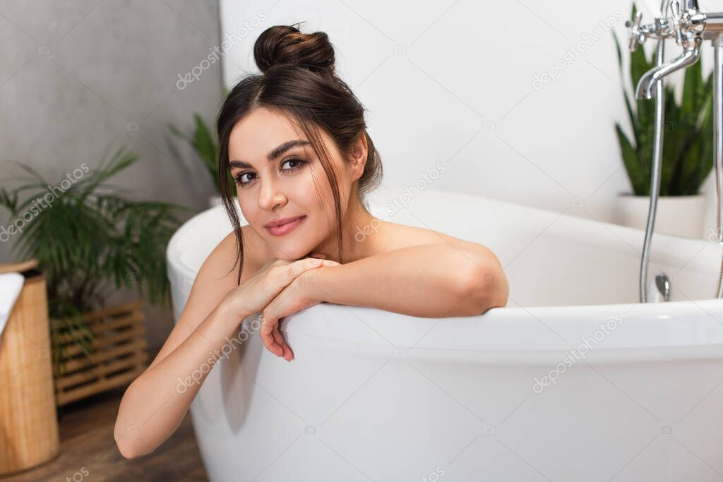 cheerful young woman with hair bun looking at camera in bathtub 